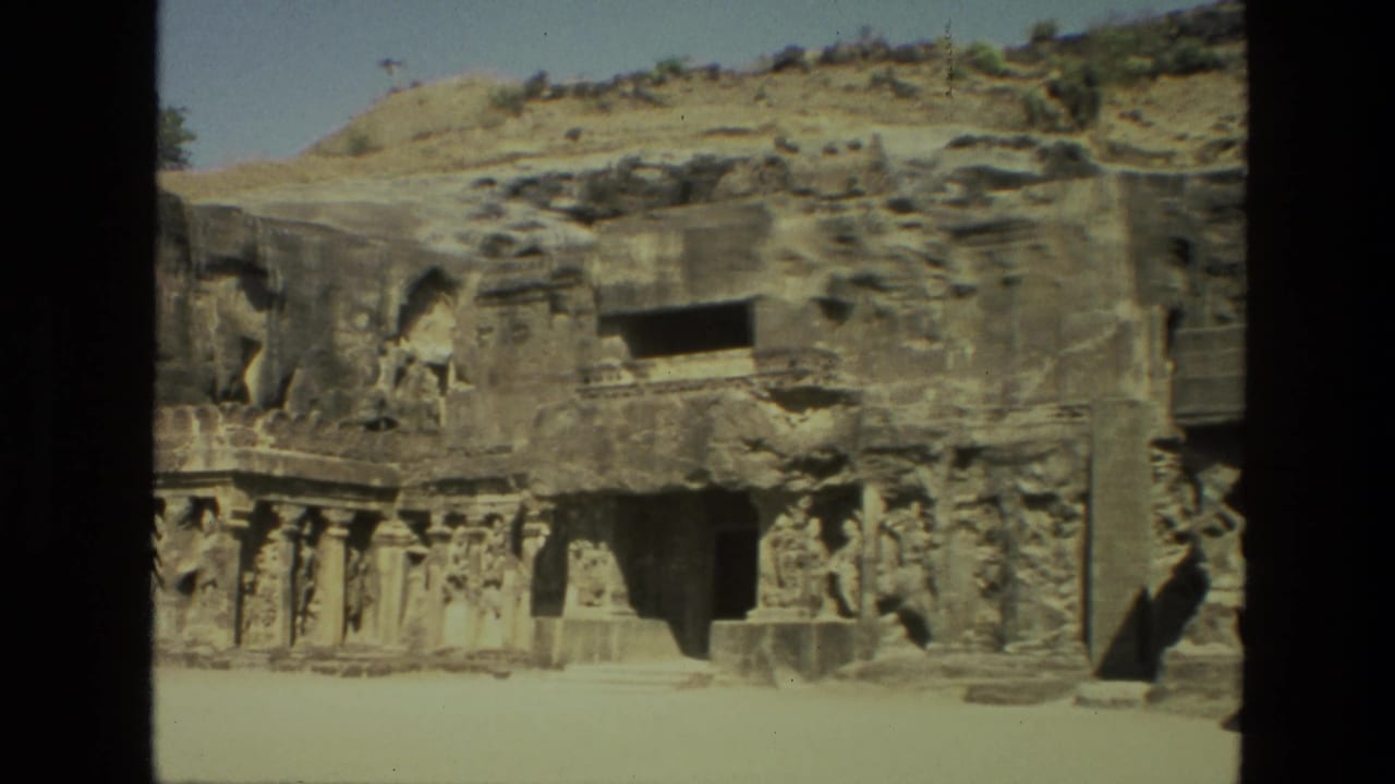 1974 India 8mm frame with a wide view of Ellora Caves carvings, columned stonework, shadowed entrances, and dry hillside above.