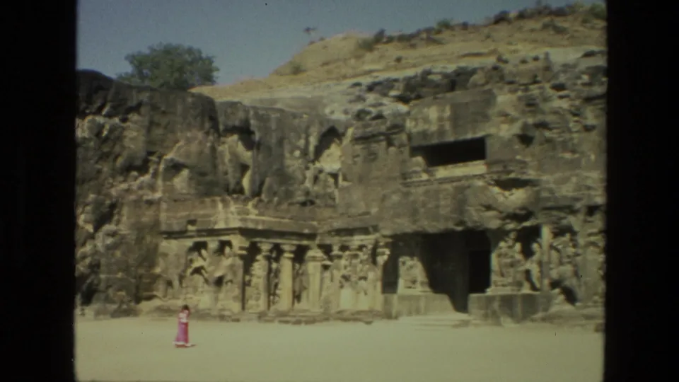 1974 India 8mm frame showing Ellora Caves stone carvings and a distant tourist in pink crossing the open courtyard.