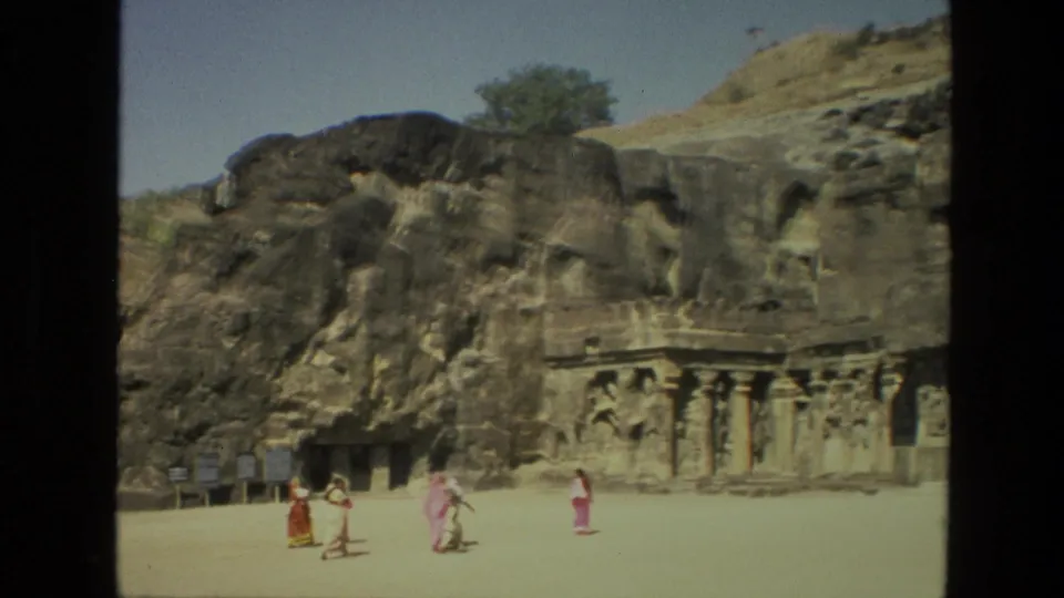 1974 India 8mm frame showing several tourists in traditional clothing walking before Ellora Caves rock-cut carvings and signboards.