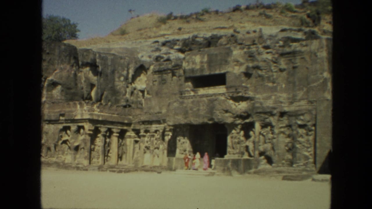 1974 8mm view of the Ajanta Caves in India, with visitors in colorful sarees near carved rock-cut entrances.