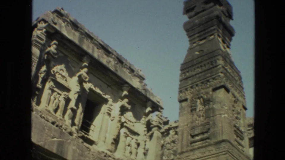 1974 8mm frame of Ajanta cave carvings with a tall stone structure at the right edge and a dark central doorway.