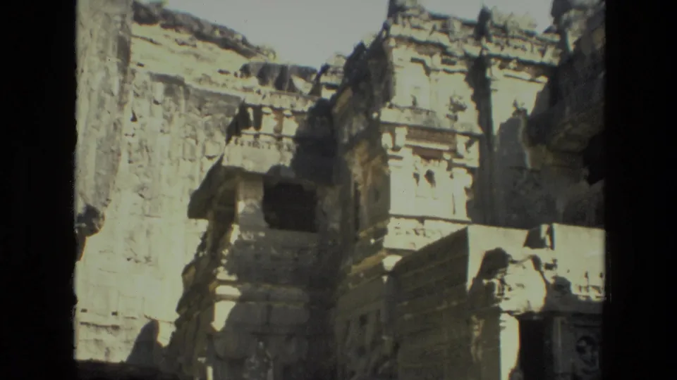 Handheld 8mm frame of a carved cliffside ruin in India, showing a projecting stone section, weathered walls, and dark recesses.