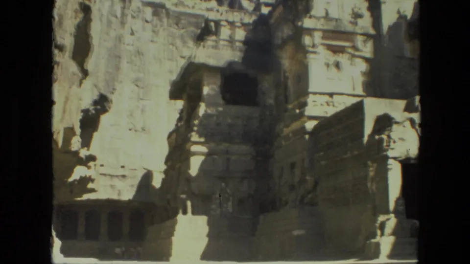 1974 India 8mm frame showing a weathered cliff-carved temple facade, stone steps, carved details, and harsh sunlight.
