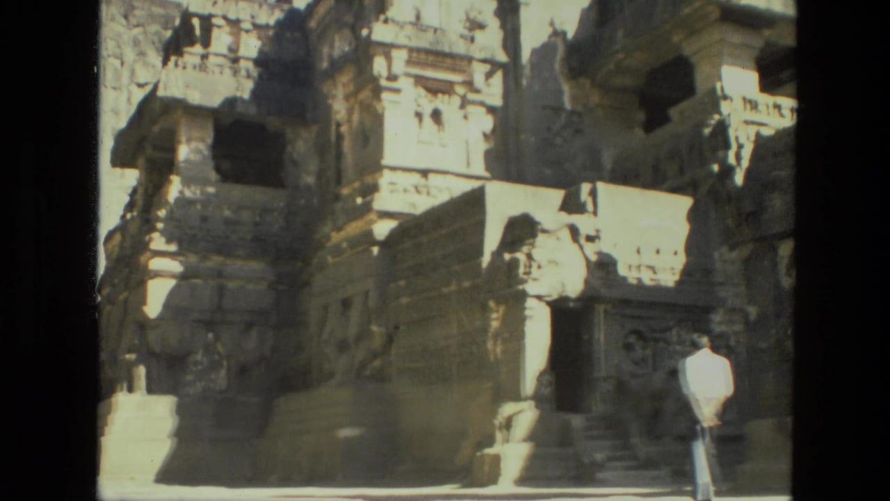 1974 India 8mm frame with temple ruins, carved stonework, rocky cliff, and a distant visitor in light clothing near the steps.