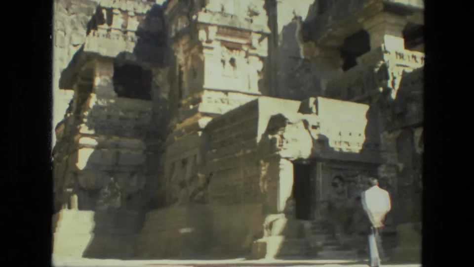 1974 India 8mm frame with temple ruins, carved stonework, rocky cliff, and a distant visitor in light clothing near the steps.