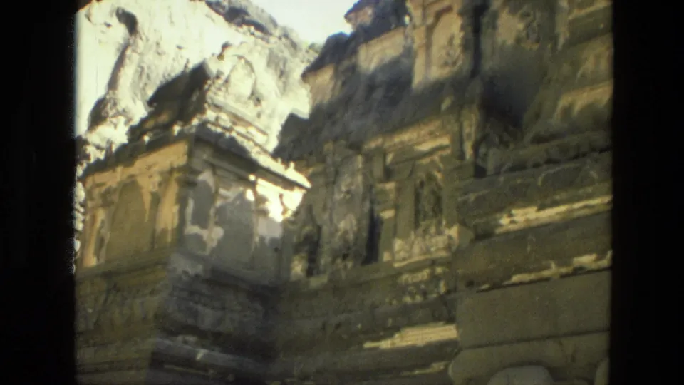 1974 India 8mm frame looking upward at cliff-carved temple architecture, with weathered stone surfaces and deep shadow.