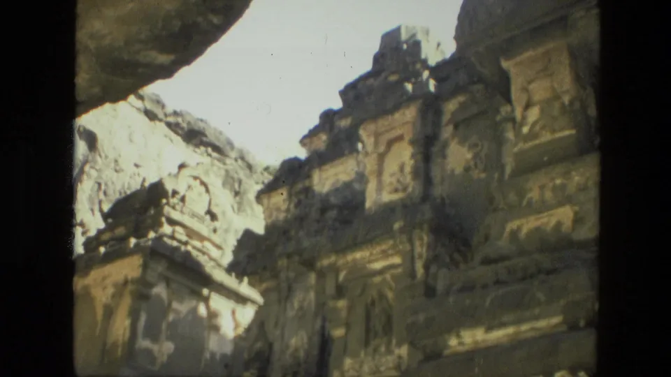 1974 India 8mm frame showing cliffside temple ruins, carved architectural panels, a rock overhang, and bright open sky.