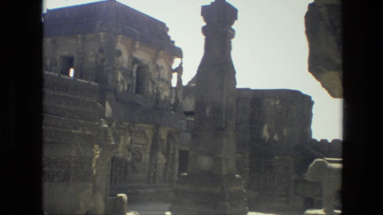 Handheld 1974 India footage frames a sunlit ruin courtyard with carved walls and a tall stone architectural element.