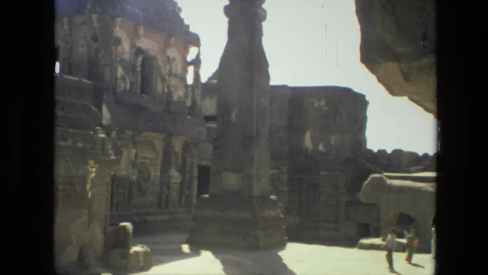 1974 home-movie frame from India showing tourists crossing a bright courtyard beside carved ancient stone structures.
