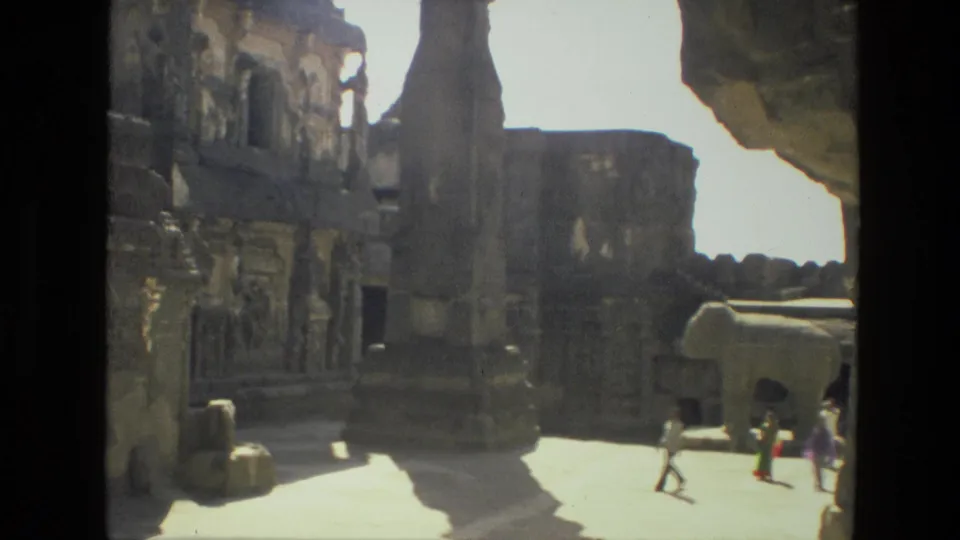 Tourists in 1974 India move through a bright ruin courtyard bordered by carved stone walls and shadowed openings.