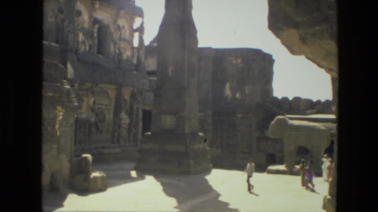 Sunlit stone courtyard with carved structures, a tall pillar, and visitors walking near the center.