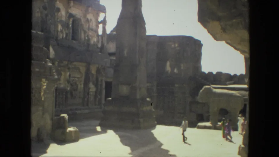 1974 India 8mm view of a sunlit stone courtyard with carved architecture, a tall pillar, and small visitor figures.