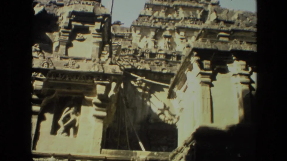 1974 India home-movie frame showing a sunlit rock-cut temple facade, dark interior opening, pillars, and carved upper details.