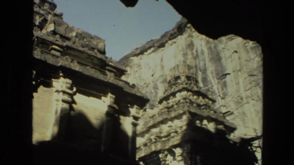 1974 India home-movie frame with a carved temple section, pale worn wall, dark foreground border, and strong sunlight on stone surfaces.
