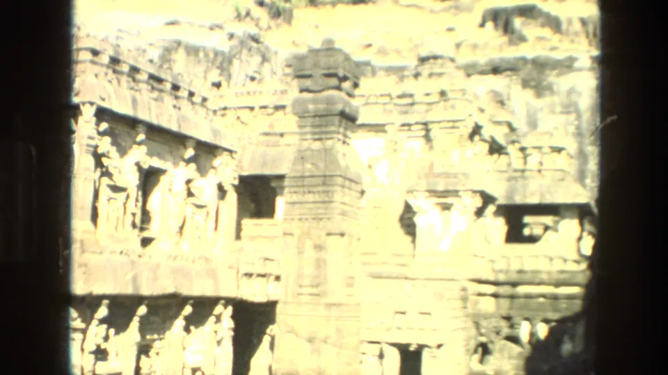 Bright 1974 India home-movie frame showing carved temple reliefs, sculptural figures, recessed openings, and a central stone pillar.