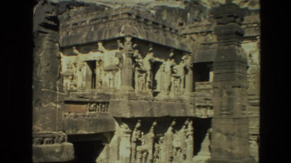 1974 India 8mm frame showing close architectural detail of a rock-cut temple with sculptural figures, ledges, and a dark doorway.