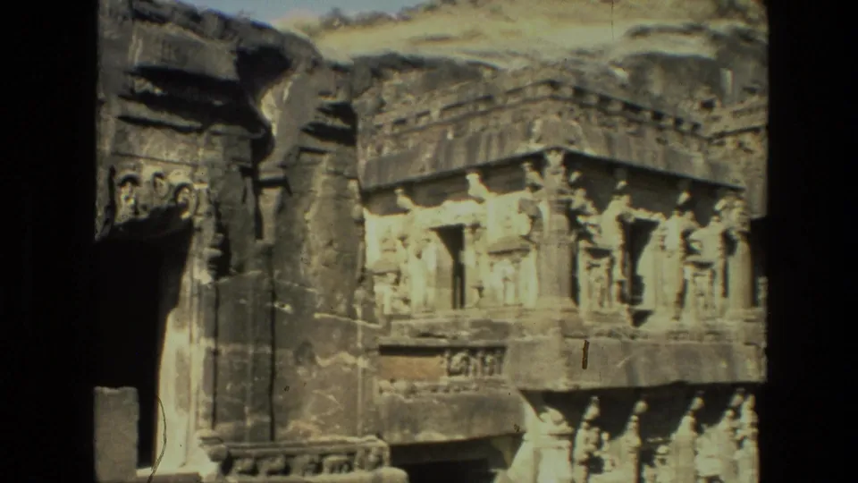 1974 India 8mm frame showing a temple facade corner with sculptural carvings, stepped stone ledges, and a dark rock-cut recess.