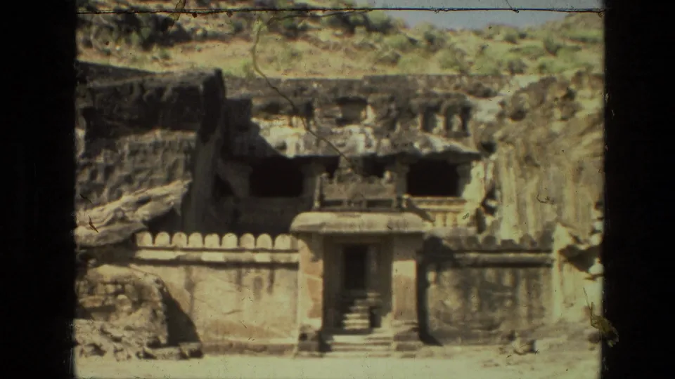 1974 India 8mm frame showing a small temple front, steps, low boundary wall, and cave openings in a rocky hillside.