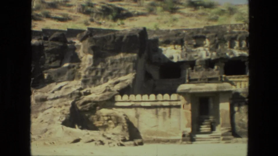 1974 India 8mm frame of a rock-cut temple area with a small entrance structure, low wall, cave openings, and hillside stonework.