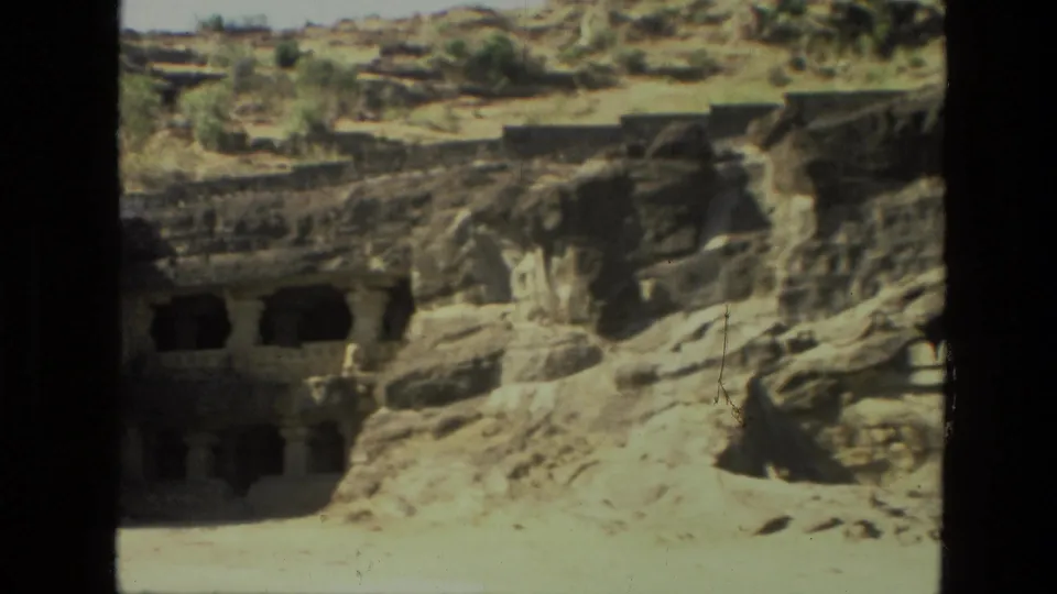 1974 India travel frame showing dark cave openings and carved stone forms set into a sunlit rocky slope.