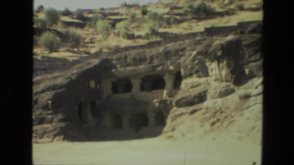 1974 8mm India frame showing a rock-cut structure with columns, shadowed entrances, and pale open ground.
