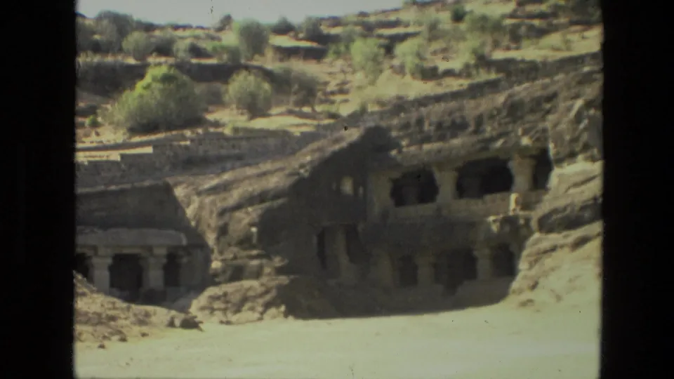 1974 India frame showing multiple rock-cut structures, dark cave openings, and a sandy foreground below a rocky hill.