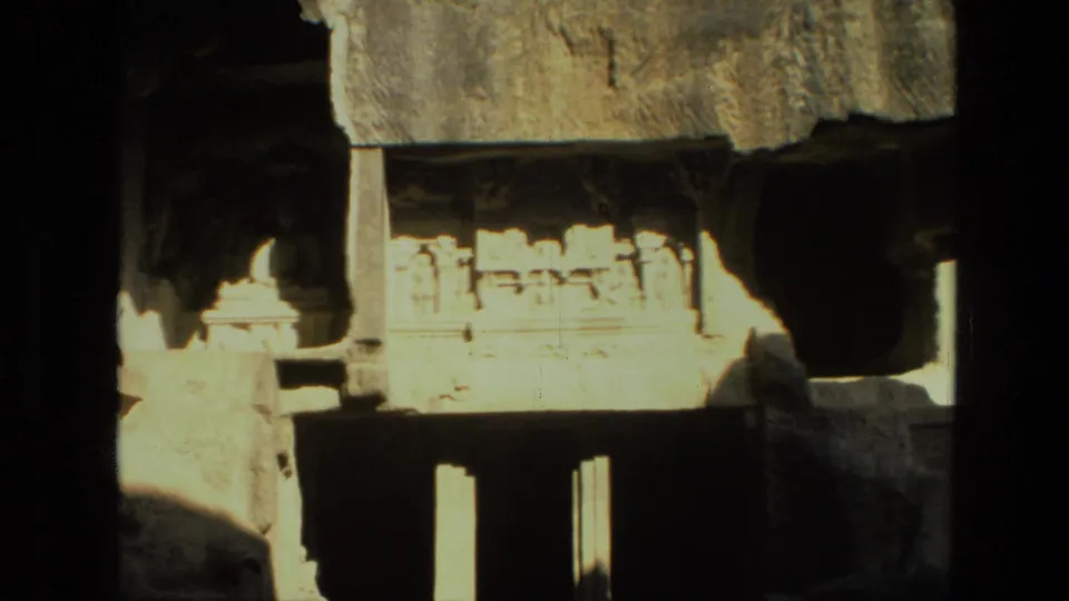 1974 8mm frame showing a bright carved stone facade in India, with shrine-like detail and dark rectangular openings beneath.