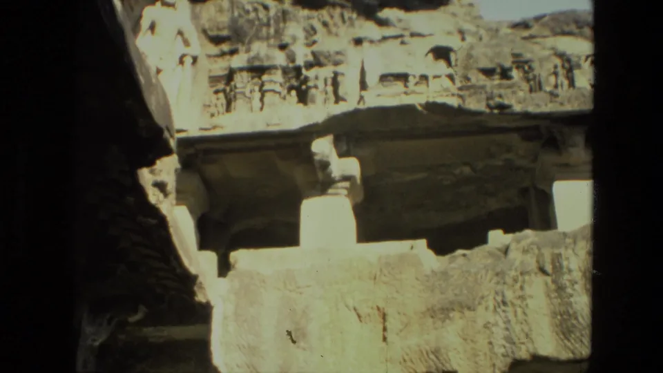 1974 India 8mm view of a carved rock-cut facade, with small recesses and a broad stone ledge in harsh light.