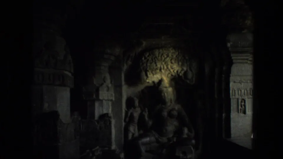 1974 8mm India frame of a shadowed cave temple interior with a seated religious statue, carved alcove, and decorated pillars.