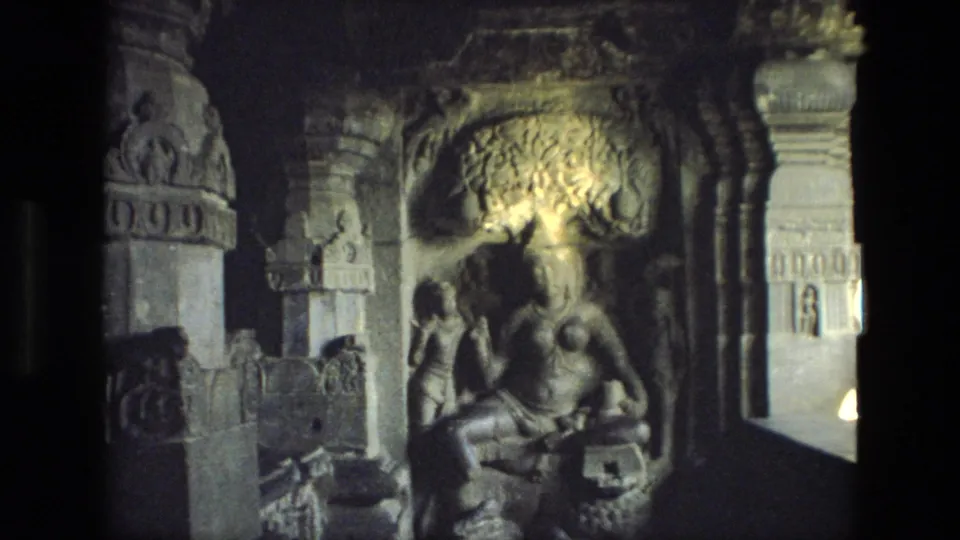 1974 8mm India travel frame showing carved temple pillars, relief surfaces, and a seated religious statue in a cave interior.