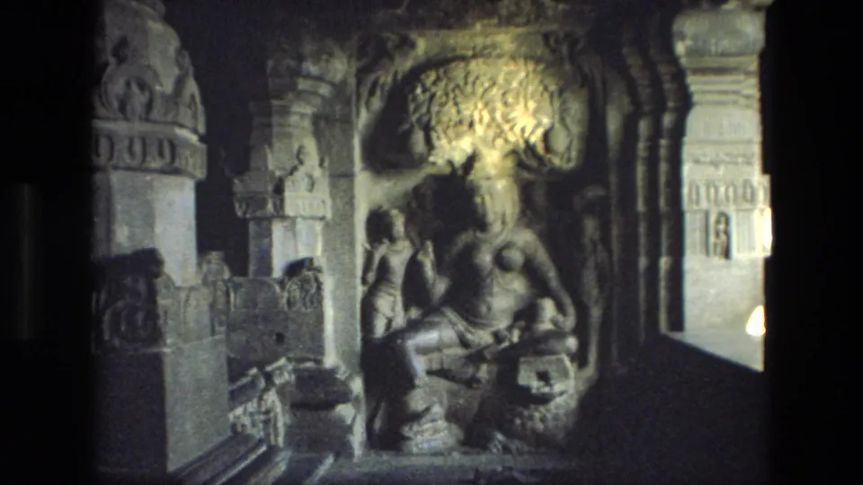 1974 8mm India frame with a seated religious statue, carved stone surround, decorated pillar, and bright side opening in a cave temple.