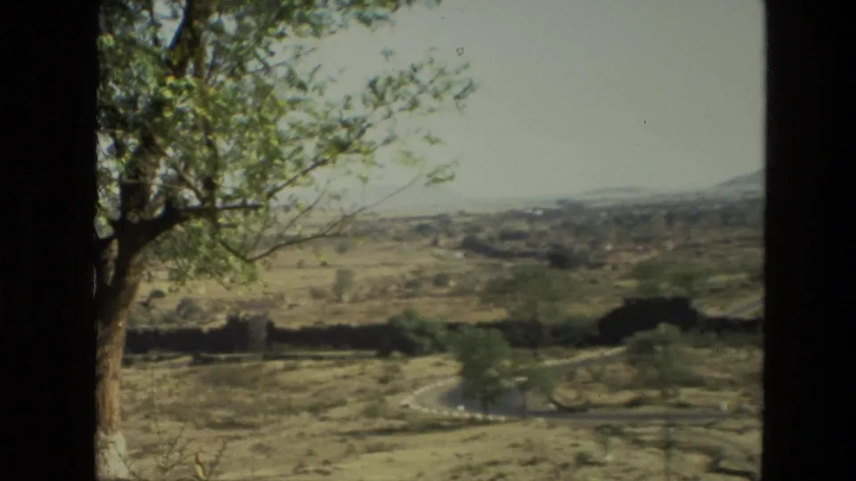 1974 India 8mm frame showing dry terrain, a foreground tree, sparse greenery, and low hills beyond the landscape.