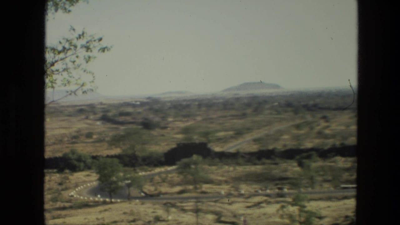 Dry rolling landscape with sparse trees, roads, and low hills seen through dark vertical framing.