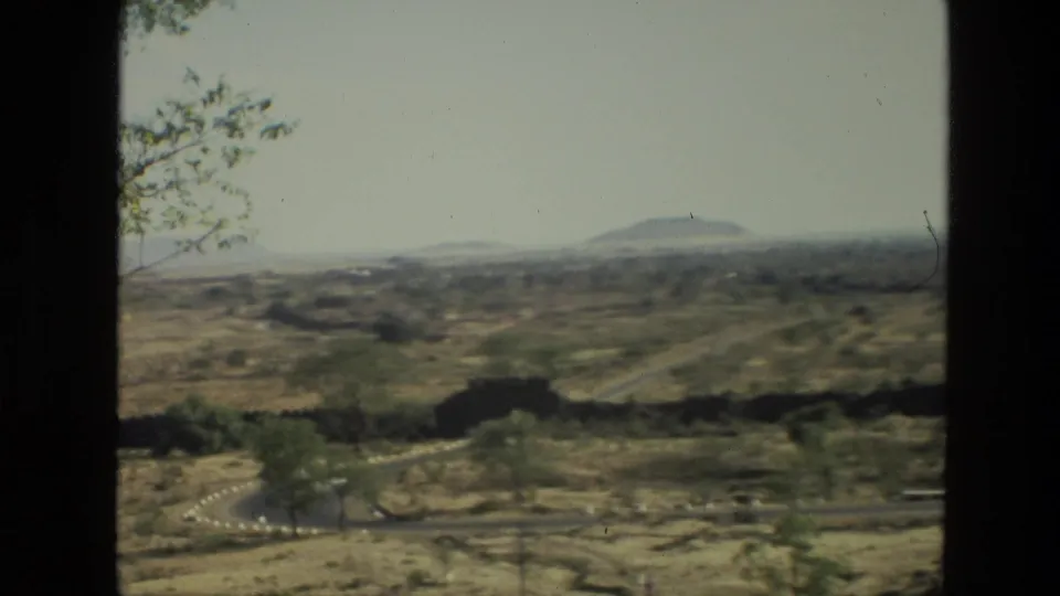 1974 India 8mm view of arid rolling countryside with scattered trees, winding roads, and distant low hills framed by dark window edges.