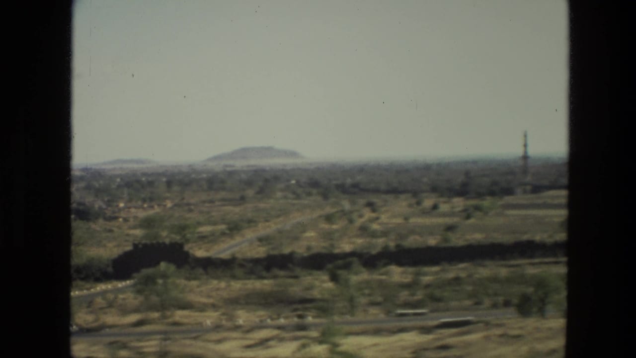 Open dry fields with a distant hill, scattered trees, road traces, and a tower near the right edge.