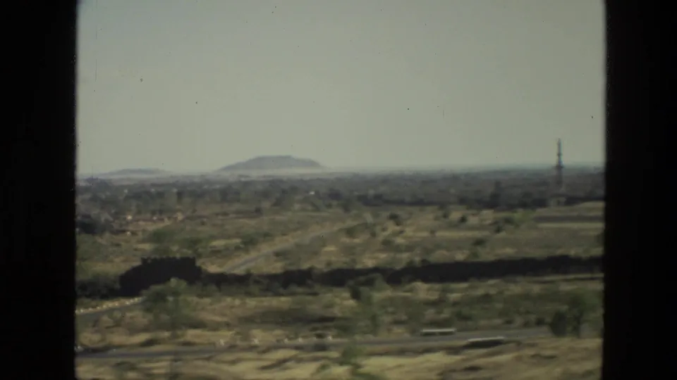 1974 India home-movie frame showing dry rural terrain, a distant rounded hill, road traces, and a slender tower near the horizon.