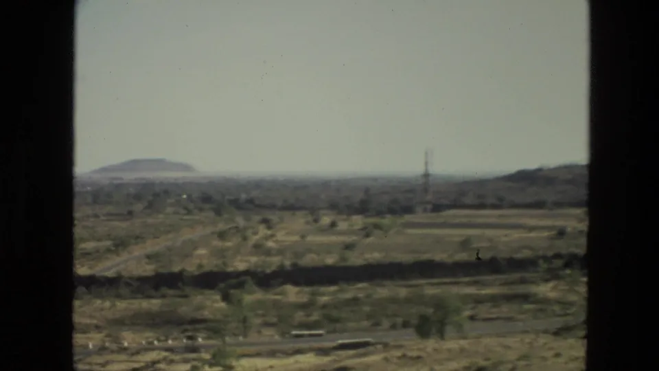 1974 India 8mm landscape view with open dry fields, a distant low hill, scattered vegetation, and road paths under a pale sky.