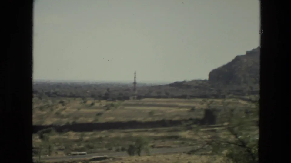 1974 India home-movie frame showing a broad dry landscape with sparse trees, a distant tower, and a rocky hillside at the right edge.