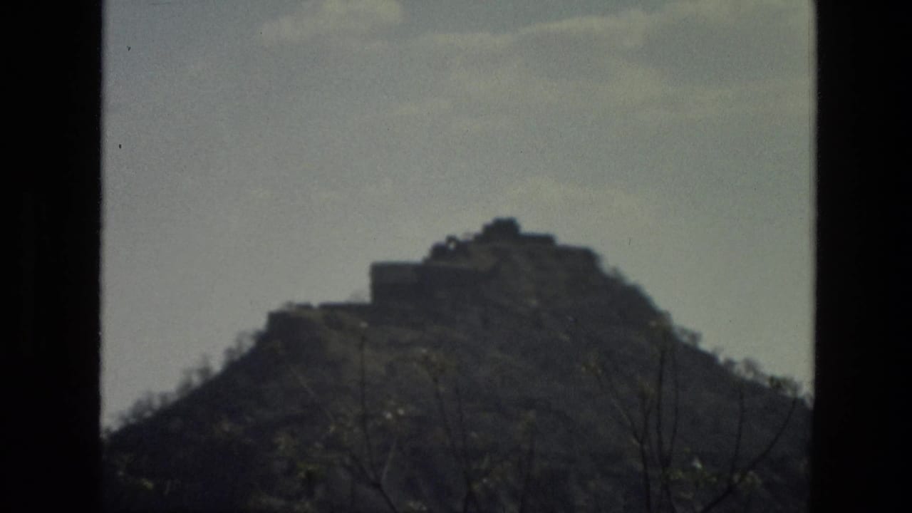 1974 India 8mm frame of a brown hill and small structure on the crest, with dark foreground framing and pale sky.