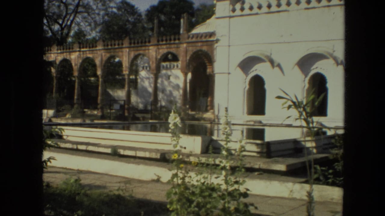 1974 India travel frame with arched colonnades, white plaster palace wall, low courtyard walls, and greenery in strong daylight.