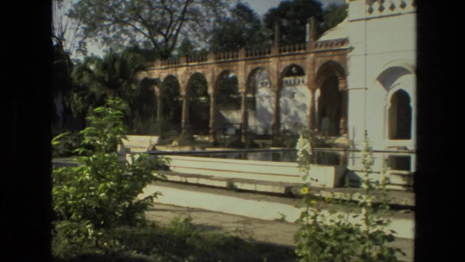 1974 India 8mm frame showing the palace courtyard from a visitor viewpoint, with arches, pool edge, shrubs, and tree shade.