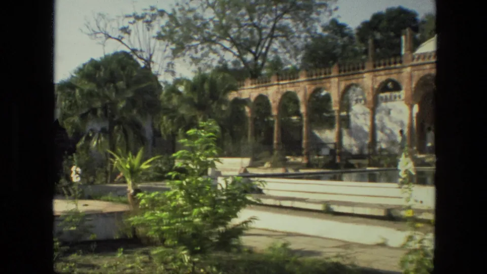 1974 India home-movie frame with palms and shrubs in front of an arched palace colonnade and courtyard water feature.