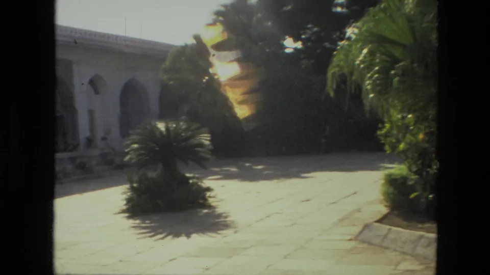 1974 India 8mm frame showing a white arched building, stone paving, and a palm shrub in strong sun.