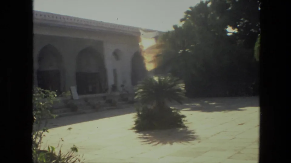 1974 India 8mm frame with a white arched exterior, palm shrub, stone paving, and bright lens flare.