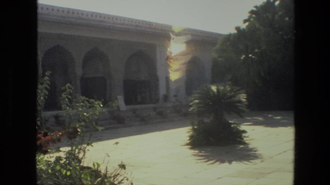Arched stone building beside a sunny courtyard with shrubs and palms.