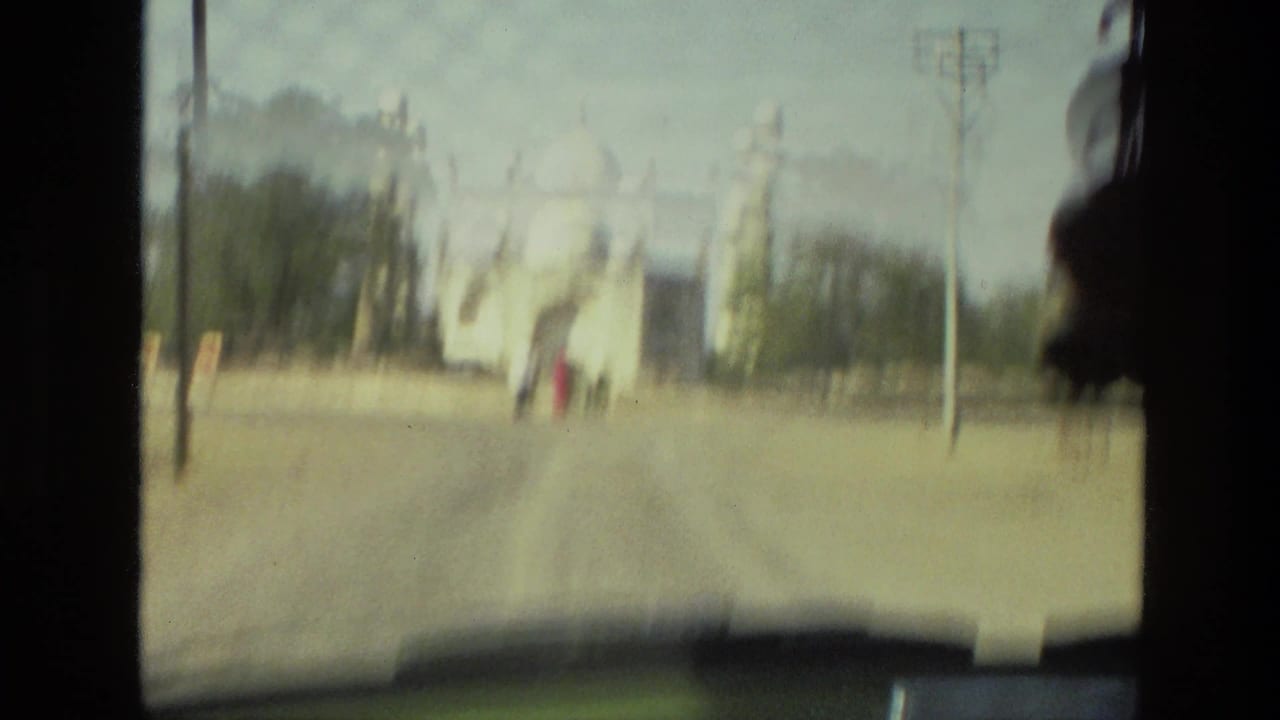 View through a vehicle windshield toward a pale monument and distant pedestrians.