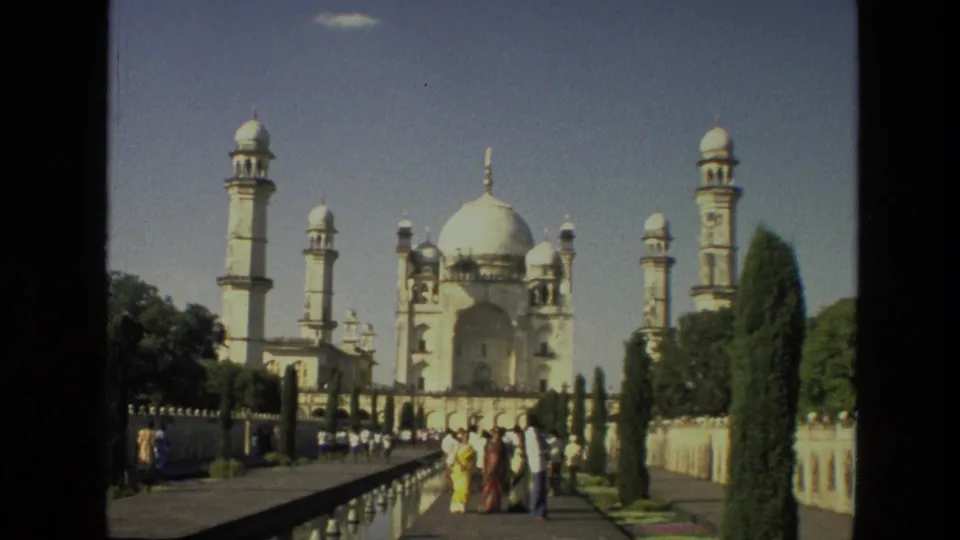Tourists on a landscaped path near the Taj Mahal, with marble domes, minarets, trees, and railings visible in 1974 footage.