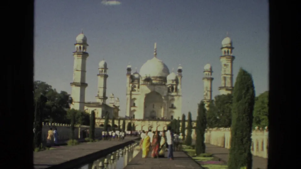 1974 India 8mm frame showing a white domed monument with minarets, a reflecting pool, garden paths, and visitors walking the grounds.