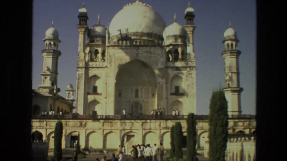 1974 India 8mm frame with a closer exterior view of the white domed religious monument, minarets, terrace wall, trees, and scattered visitors.