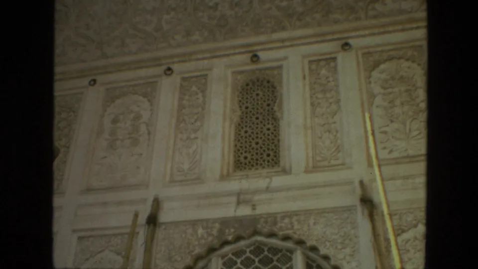 1974 India home-movie frame showing pale carved temple wall panels, vertical dark frame edges, lattice inserts, and part of an arched window below.